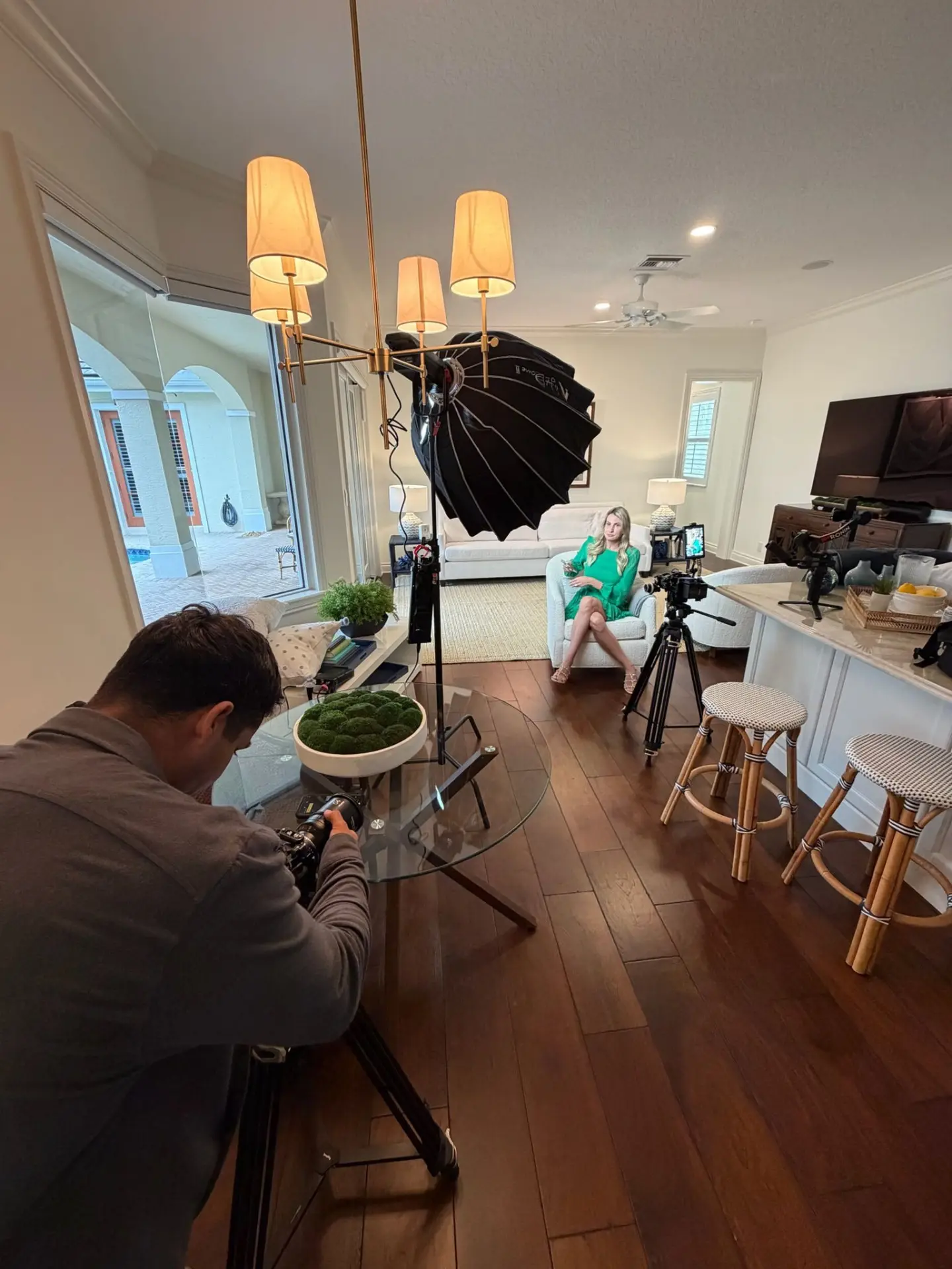 Behind the scenes: a camera operator films an interview with a seated subject in a warmly lit living room set, with pro lighting and camera gear visible.